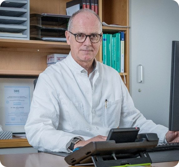 Dr. Grotkamp sitzt am Schreibtisch vor Computer und Telefon in Büro mit Regalen