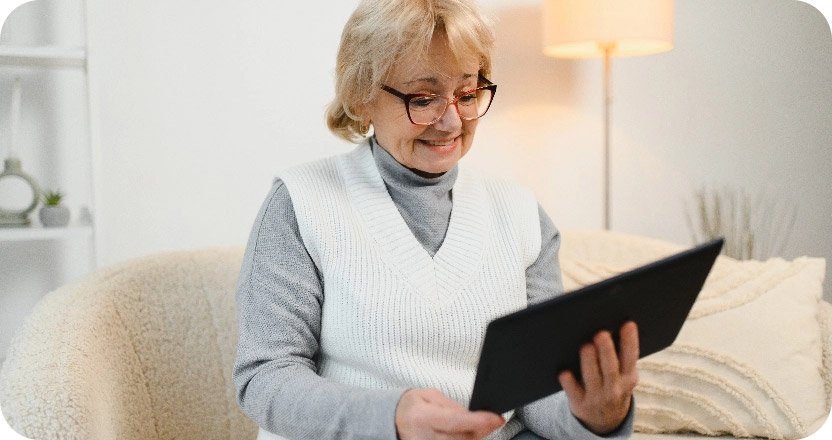 Ältere Frau mit Brille und hellgrauem Pullover sitzt auf Sofa und hält Tablet in den Händen