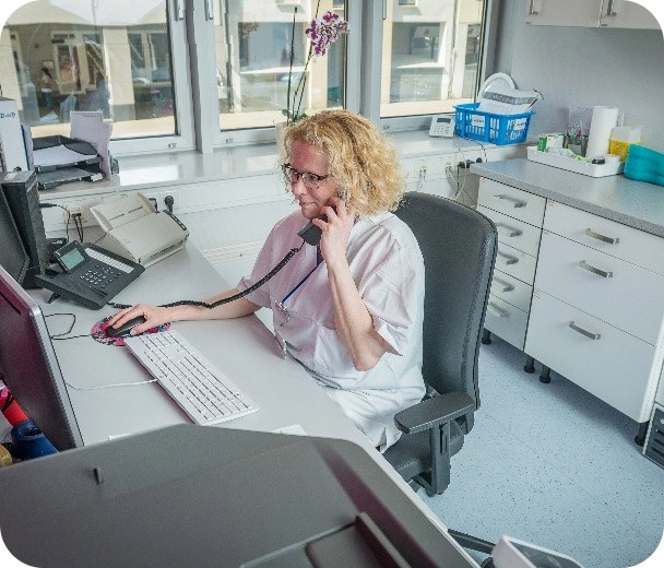Krankenschwester sitzt an einem Schreibtisch, telefoniert und bedient eine Computermaus in einem hellen Stations-Büro