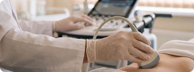 Arzthand mit Handschuh hält Ultraschallsonde auf Bauch eines liegenden Patienten vor Ultraschallgerät mit Bildschirm