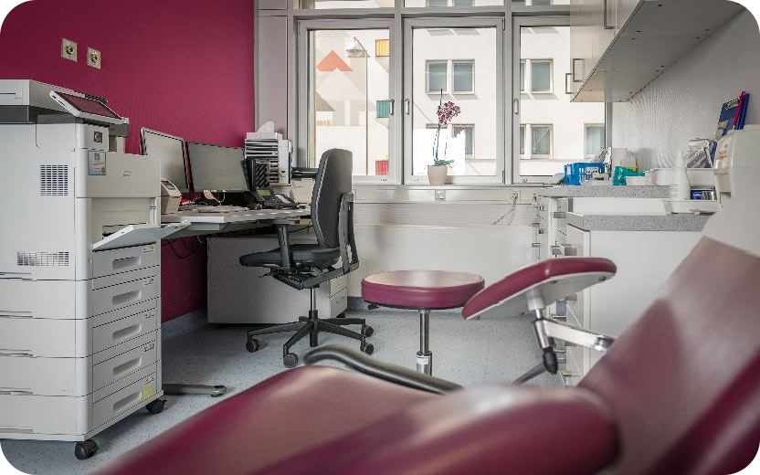 Untersuchungszimmer mit rotem Untersuchungsstuhl, Drehstuhl, Schreibtisch mit Computer und Fenster mit Blick auf Nachbargebäude