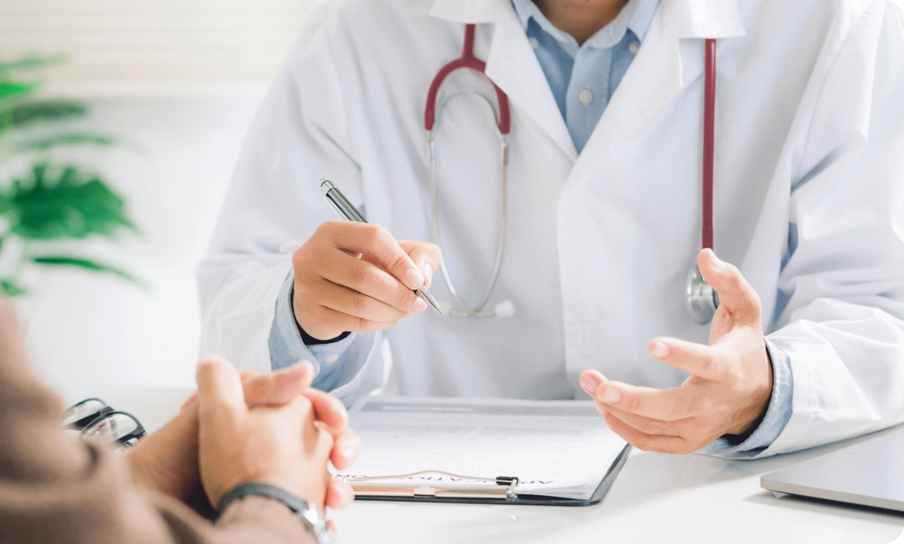 Arzt mit weißem Kittel und rotem Stethoskop erklärt einem Patienten etwas am Schreibtisch